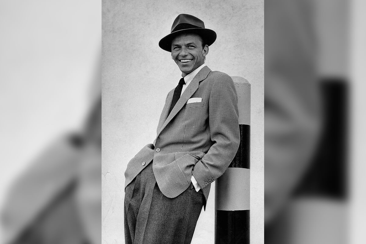 Frank Sinatra smiles at the camera while leaning against a post.